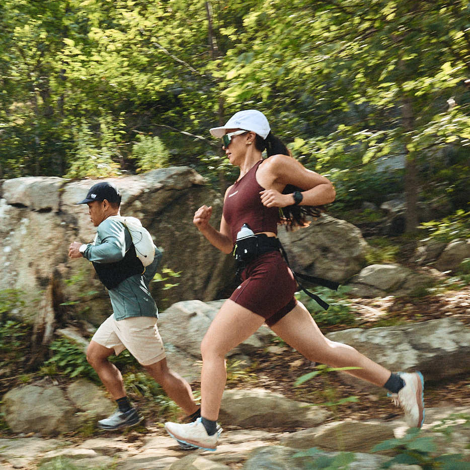 Two runners sprinting on a rocky forest trail, a woman in maroon shorts and a man in light shorts wearing hats and socks