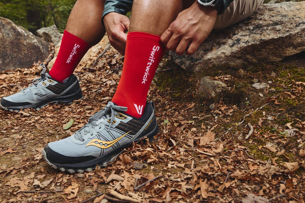 Close-up of legs wearing red Swiftwick crew socks showing logo and snug fit with gray trail shoes on leaves