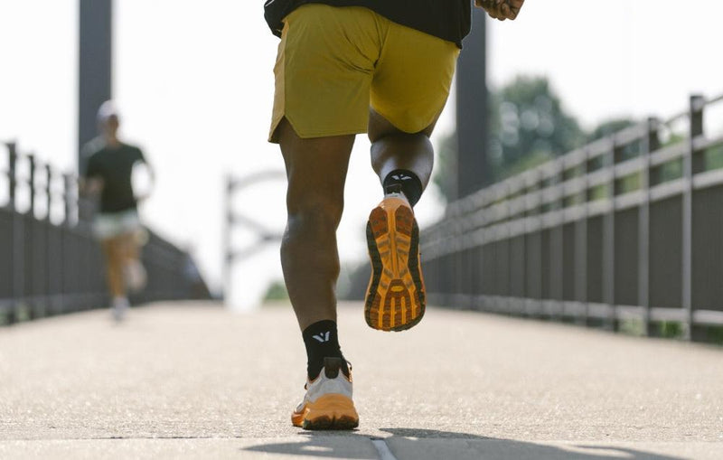 Runner wearing yellow shorts and orange-soled shoes running on a bridge, with another runner blurred in the background.