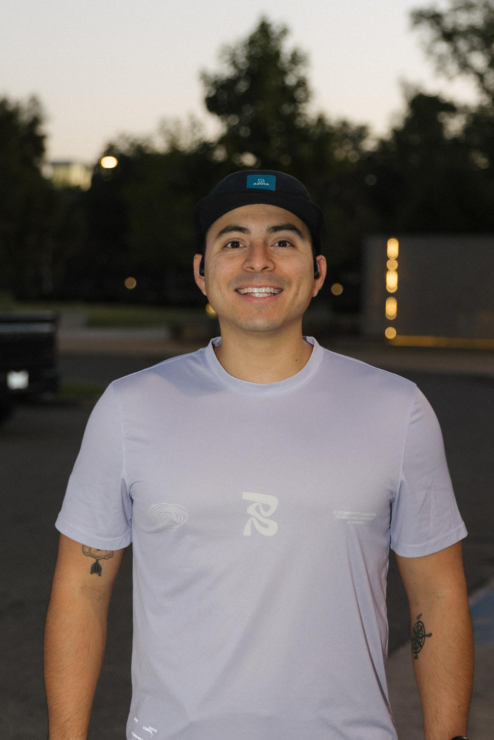 A smiling person wearing a light purple shirt and a black cap, standing outdoors.