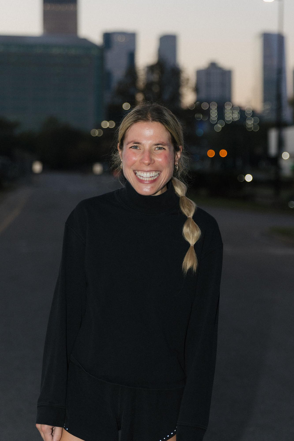 A smiling woman in a black outfit standing on a city street at dusk.