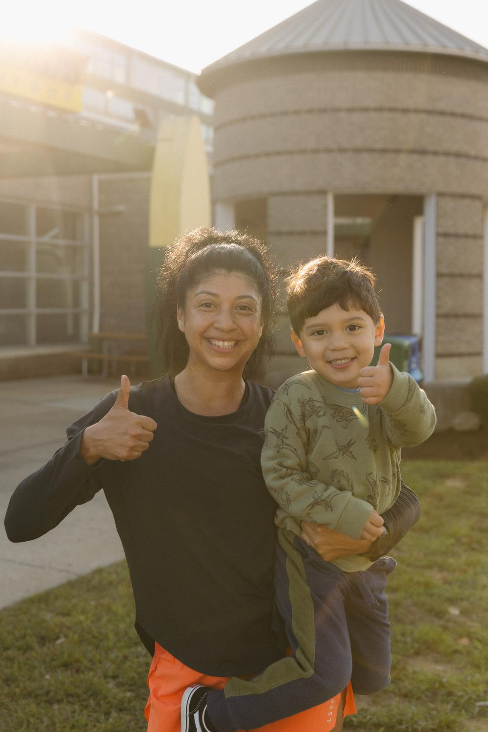 A woman holding a smiling child, both giving thumbs up outdoors.