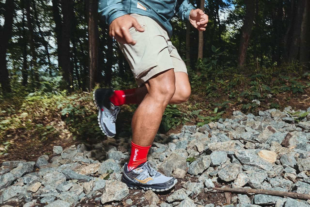 Person running on a rocky forest trail wearing red crew socks and gray trail shoes