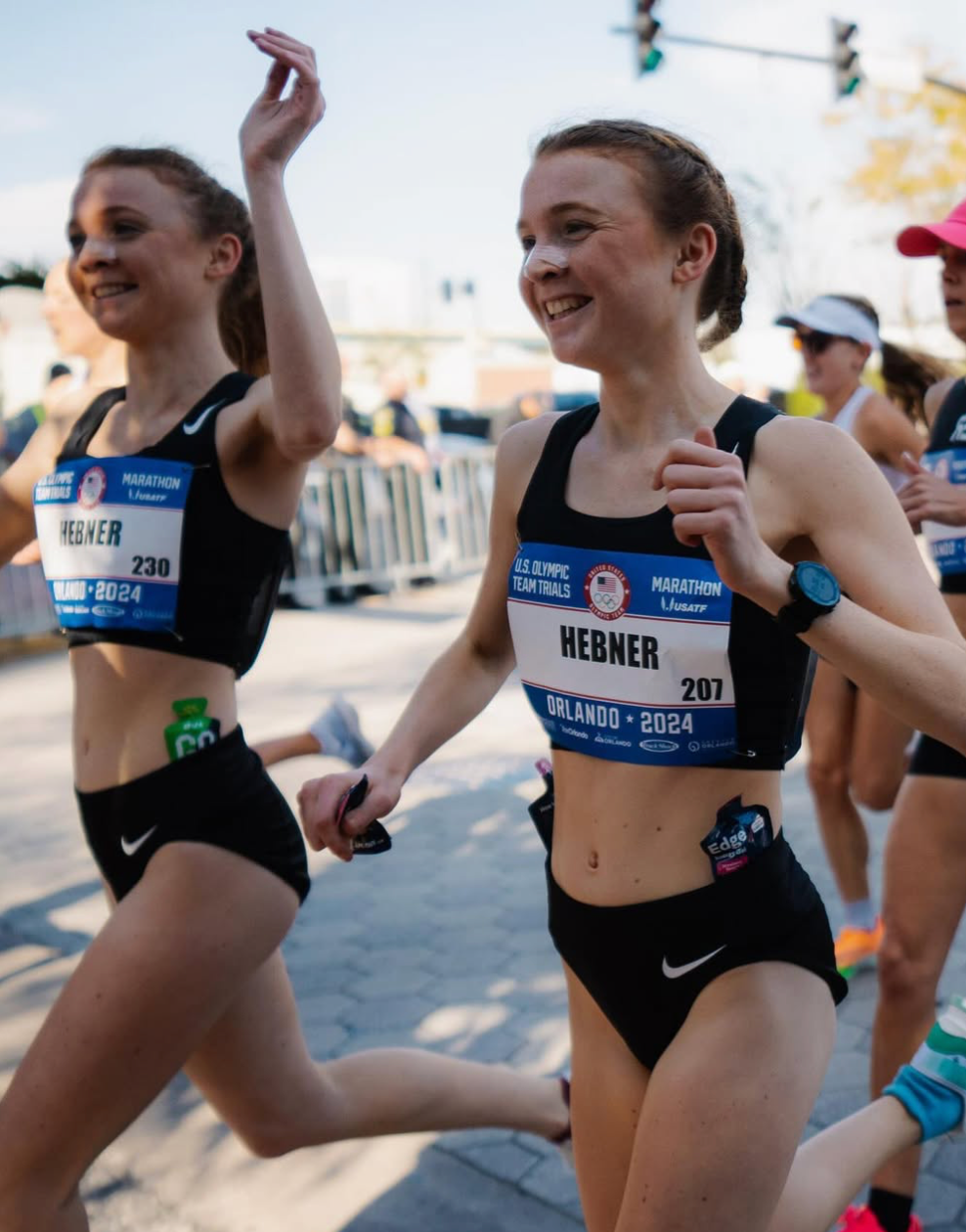 Two female marathon runners smiling while running, one with bib 'HEBNER 207'.