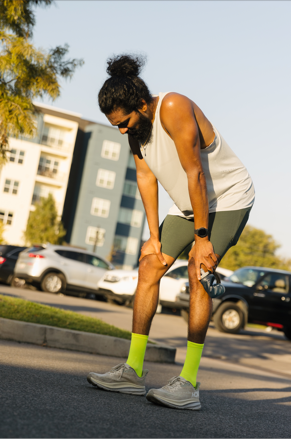 Runner bent over with hands on knees holding a bottle on a street, wearing white tank, green shorts and neon yellow socks