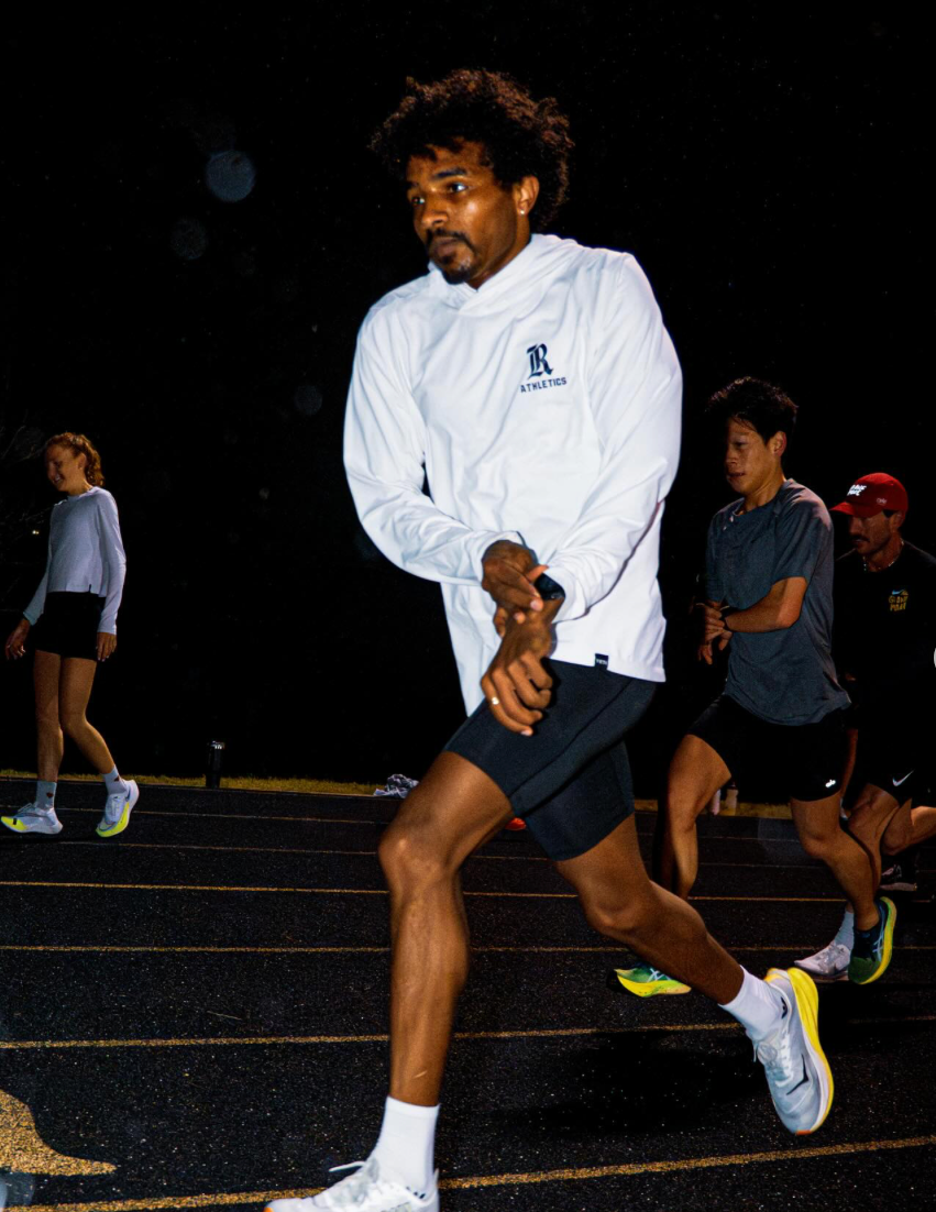 A runner in a white hoodie and shorts training on a track at night.