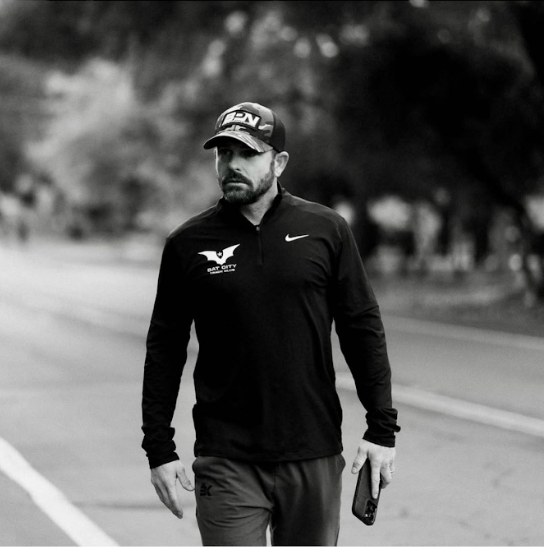 A man walking on a road wearing a black long-sleeve shirt and a cap, holding a phone.