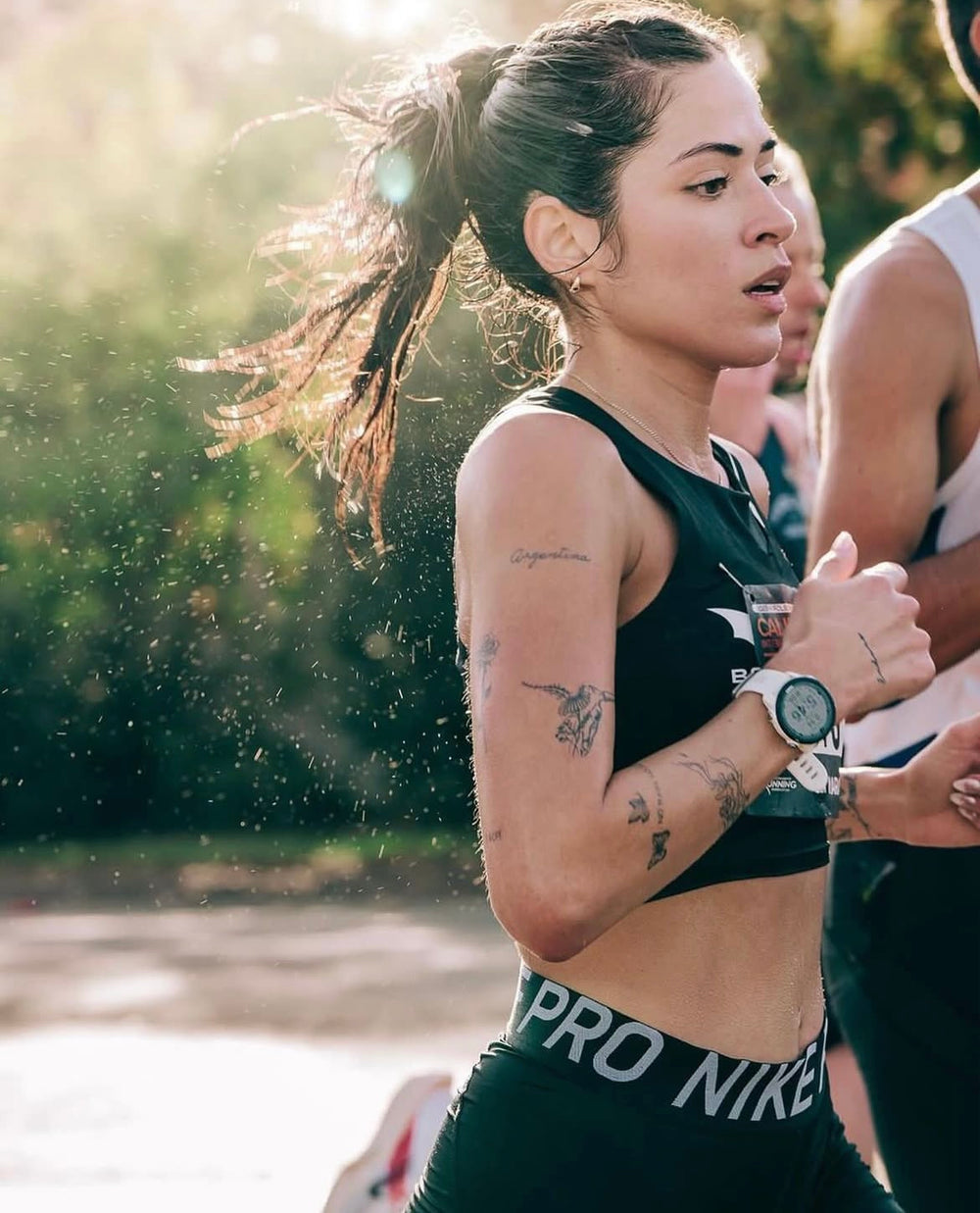A woman running in athletic wear, with tattoos and a watch, in a sunny outdoor setting.